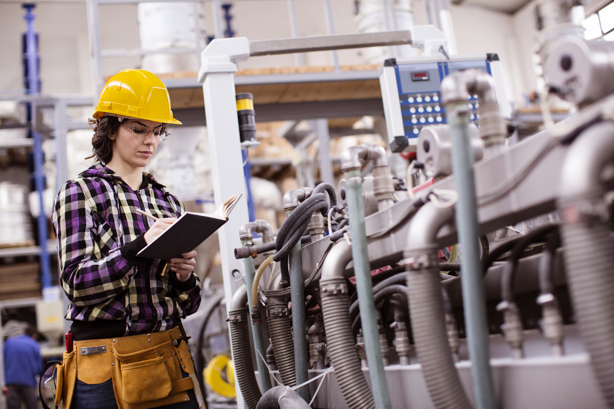 A female machine technician writing notes to learn components of a large industrial machine