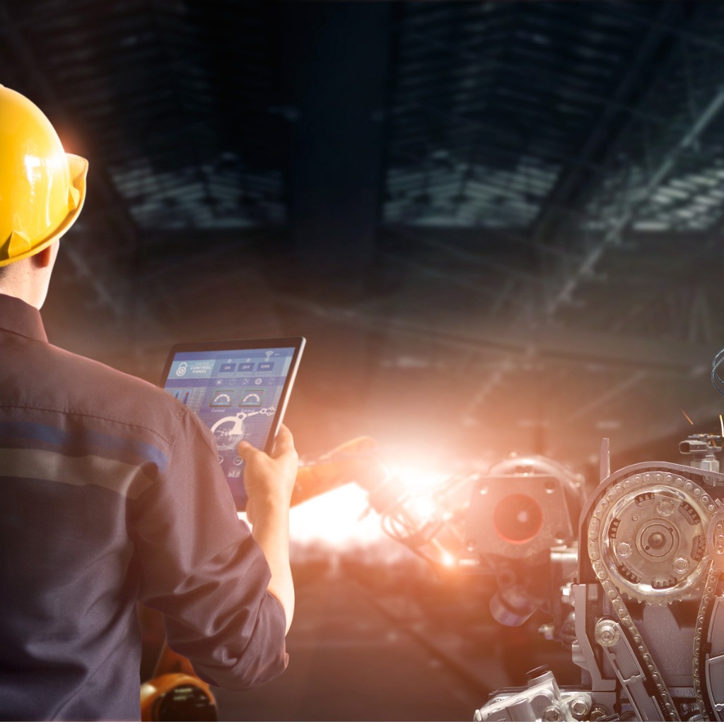 A machine technician in a yellow hardhat monitors machine performance on a tablet in a factory