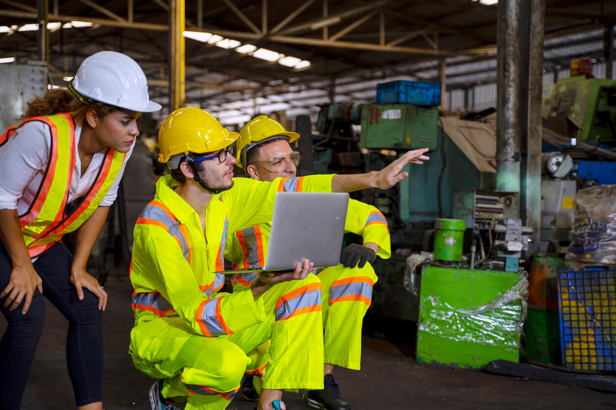 Several individuals in high-vis jumpsuits use a laptop to check nearby equipment in an industrial setting.