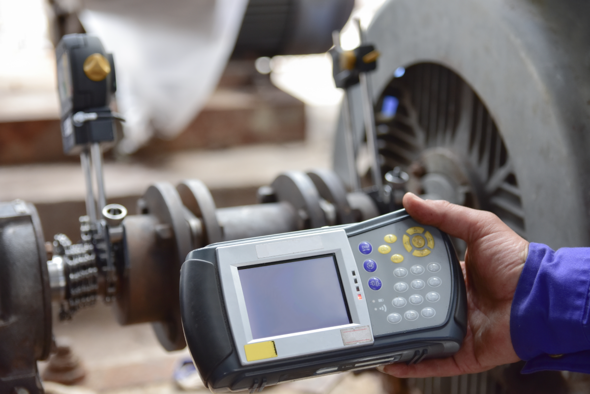 A hand holds a diagnostic device with a screen and buttons near industrial machinery. The setting suggests a technical inspection or maintenance task.