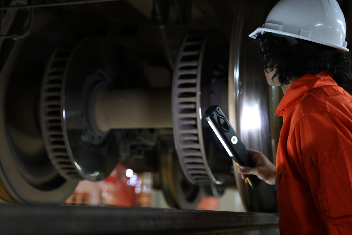 A person in a white hard hat and orange safety uniform inspects rotating equipment with a flashlight. The scene conveys focus and technical precision.