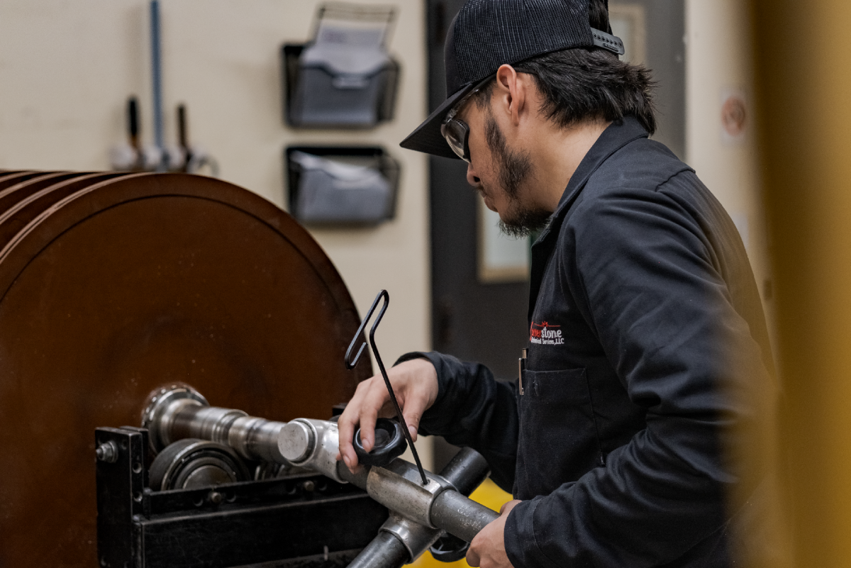 Craftsman in safety glasses operates a large metal lathe, shaping a cylindrical workpiece in a workshop; focused, hands-on mood.